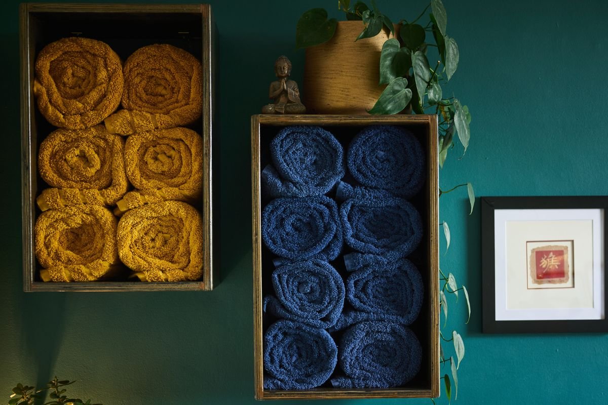 Neatly rolled mustard and blue therapy towels in rustic wooden shelves against a teal wall, showcasing the serene and professional environment of Steven Murdoch’s London clinic.